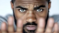 Close up portrait of young African American man with intense gaze and beard looking through blurred fingers held in front of face against light background.