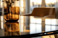 Amber glass tumbler on reflective black surface with soft window light creating atmospheric mood. Minimalist home interior with blurred furniture background.