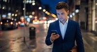 Young professional businessman using smartphone on city street at night with bokeh lights background for business communication and urban lifestyle.