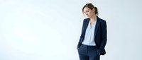 Middle-aged Caucasian woman in professional business attire, wearing navy blazer and light blue shirt, standing with hand in pocket, looking thoughtful. Corporate professional portrait.