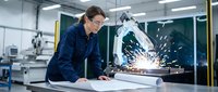 Male engineer in dark blue work shirt reviewing blueprints at industrial workstation with welding sparks visible in background manufacturing facility.