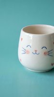 Cute white ceramic mug with cat face design featuring pink cheeks and blue whiskers on turquoise background for kitchen decor and beverage enjoyment.