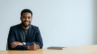 Young African American male professional in dark blazer seated at desk with notebook, smiling at camera for corporate business portrait.