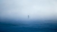 Solitary figure standing in vast misty ocean waters during foggy weather conditions. Atmospheric seascape with minimal visibility and moody blue tones.