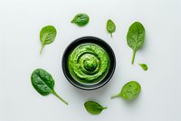 Fresh green spinach sauce in black bowl surrounded by raw baby spinach leaves on white background, overhead view for food photography and menu design.
