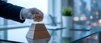 Close-up of businessman's hand placing wooden block with dollar sign on pyramid stack, symbolizing financial growth and wealth building strategy in modern office setting.