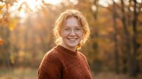 Happy young Caucasian woman with curly blonde hair wearing cozy brown sweater smiling outdoors in autumn park with golden sunlight and colorful fall foliage.