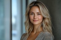 Young Caucasian woman with wavy blonde hair and natural makeup smiling confidently at camera. Soft window lighting creates professional headshot with blurred background.