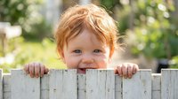 Adorable toddler with curly red hair peeking over wooden fence in sunny backyard, smiling at camera with bright blue eyes and cheerful expression.