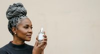 Woman with braided updo hairstyle holding clear fragrance bottle against neutral beige background, suitable for beauty, perfume, and cosmetics marketing campaigns.