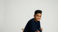 Young man with dark hair and olive complexion wearing navy blue casual shirt, seated against white background, looking over shoulder with confident expression, isolated for design use.
