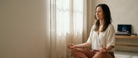 Young woman practicing mindfulness meditation in bright modern bedroom sitting cross-legged on floor wearing comfortable white clothing for wellness lifestyle.
