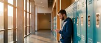 Young male student using smartphone in modern school hallway with lockers during golden hour sunlight streaming through windows.