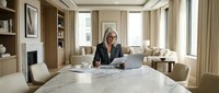 Mature woman with glasses working on laptop at marble table in modern luxury home office with built-in shelving and fireplace.