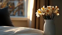 Fresh daffodils in white ceramic vase on bedside table catching morning sunlight through window, creating cozy spring atmosphere in bedroom interior.