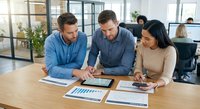 Three diverse business professionals collaborating over financial charts and data analysis in modern office workspace for strategic planning meeting.
