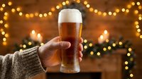 Unrecognizable person in cozy sweater holding cold beer glass with foam head against warm bokeh lights and festive garland backdrop for holiday celebrations.