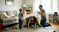 Happy family playing together at home with father holding blanket over mother and children in cozy living room during quality time bonding activity.