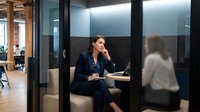 Middle-aged woman with dark hair and olive complexion, wearing navy blazer, sitting in modern glass-walled office meeting room, hand near chin in thoughtful pose, engaged in professional discussion or interview with colleague.