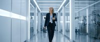 Blonde woman in dark business suit and lanyard walking through modern corporate hallway with glass offices and bright lighting, suitable for business, technology, and professional workplace concepts.
