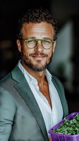 Confident mature businessman with curly hair and green glasses smiling warmly while holding fresh microgreens, wearing casual blazer and white shirt.