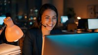 Happy young businesswoman with headset working late at night in modern office, smiling while using laptop computer for customer service or remote work.