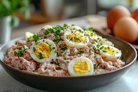 Creamy pink tuna salad with sliced boiled eggs, fresh herbs, and mayonnaise dressing served in dark ceramic bowl. Fresh ingredients for healthy lunch.