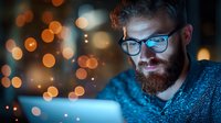Young bearded man in glasses working on laptop at night with glowing bokeh lights creating atmospheric mood for remote work and technology concept.