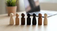 Diverse wooden peg people figures in various skin tones arranged in row on office desk representing equality, inclusion and teamwork in workplace.