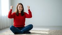 Young Asian woman in red sweater celebrating success with raised fists while sitting cross-legged on carpet at home, expressing joy and achievement.