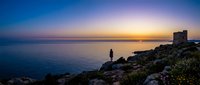 Solitary figure standing on rocky coastline at sunset, watching golden light reflect across calm sea with ancient ruins visible on distant cliff.