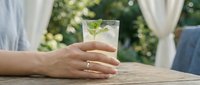 Woman's hand with wedding ring holding glass of mojito cocktail on wooden table outdoors with garden backdrop, suitable for beverage, lifestyle and celebration content.