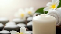 Spa wellness scene with white candle, plumeria flowers and massage stones creating peaceful relaxation atmosphere for meditation and aromatherapy treatments.