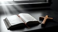 Open bible with wooden cross on dark table illuminated by dramatic sunlight rays streaming through window creating spiritual atmosphere for worship and prayer.