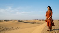 Woman in rust-colored dress standing on sand dune in vast desert landscape under clear blue sky, suitable for travel and lifestyle editorial use.