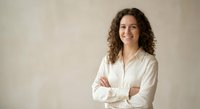 Confident young Hispanic businesswoman with curly hair smiling with crossed arms in professional white shirt against neutral background for corporate use.