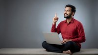 Young Middle Eastern businessman in red shirt sitting cross-legged with laptop pointing up having eureka moment against gray background.
