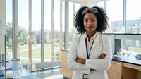 Young woman with dark curly hair and medium brown skin tone wearing professional white blazer with ID badge in modern office with floor-to-ceiling windows overlooking cityscape. Corporate professional portrait.