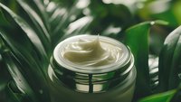 Luxurious white cosmetic cream in glass jar surrounded by fresh green tropical leaves, natural skincare product photography with soft focus effect.