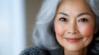 Elegant mature Asian woman with silver gray hair smiling warmly at camera in soft natural light for lifestyle and wellness content.