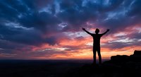 Male silhouette with raised arms celebrating success against dramatic sunset sky with purple and orange clouds over mountain landscape for motivation concepts.