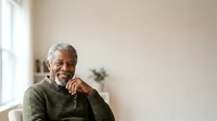 Senior African American man with gray hair wearing olive green sweater, sitting thoughtfully by window with natural light, suitable for healthcare, wellness, and lifestyle editorial use.