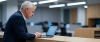Senior male professional with gray hair, Caucasian ethnicity, wearing dark business suit, working on laptop at wooden desk in modern office environment with blurred background.