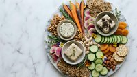 Healthy appetizer platter with hummus, fresh vegetables, crackers and nuts arranged on marble surface for entertaining and snacking.
