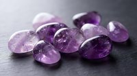Collection of polished amethyst gemstones in various purple shades scattered on dark surface for jewelry making and crystal healing practices.