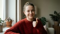 Young Caucasian woman in red knit sweater giving thumbs up gesture while sitting by window in cozy home interior with plants.