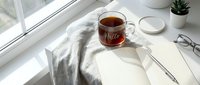 Cozy morning workspace with a glass cup of tea, open notebook, pen, and glasses on a white windowsill in soft natural light.