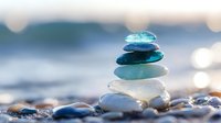 Balanced stack of smooth sea glass stones on beach pebbles with soft ocean waves and sunlight bokeh creating peaceful zen meditation atmosphere.