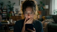 Young woman of mixed ethnicity with long wavy hair, wearing dark clothing, holding smartphone and touching her face with surprised expression in modern living room with plants and furniture.