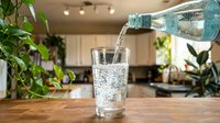 Fresh sparkling water being poured from bottle into glass on wooden kitchen counter with green plants creating healthy hydration lifestyle scene.